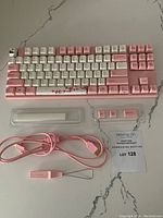 Full view of pink Varmilo keyboard with white and pink keycaps including cherry blossom spacebar, pink USB cable, keycap puller tool, and extra pink keycaps in a transparent case laid on a white marble surface.