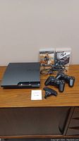 Sony PS3 CECH-2001A console with two wireless controllers, wireless keypad, and two PS3 game cases on wooden surface.
