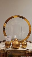Front view of the signed Annieglass glass serving bowl with wide gold rim, displayed with three gold faux fruit pieces resembling two apples and one pear on a reflective surface.