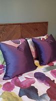 Pair of dark purple silk throw cushions set on a bed with a floral duvet cover, showing size and texture.