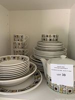 Full set of dinnerware stacked and displayed on shelf including plates, bowls, teapot, cups, and saucers with geometric mid-century modern pattern.