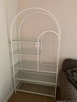 Front view of the white metal etagere shelf unit showing arched frame and glass shelves.