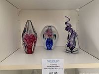 Three solid art glass sculptures displayed side by side on a white shelf, showing their varying shapes and internal colorful designs.