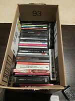 Top-down photo showing a cardboard box filled with approximately 35 music CDs stacked vertically, showing various album spines with different colors and artist names.