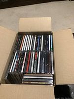 Open cardboard box filled with approximately 45 rock CDs stacked vertically revealing spines with artist names and album titles.