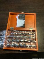 Top-down view of Mastercraft wood drill bits in sealed plastic wrappers inside an orange wooden box, showing assorted sizes and the fabric insert at the lid.