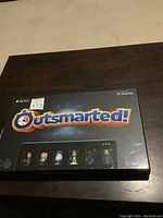 Front view of Outsmarted! board game box sealed, showing logo and character pieces inside.