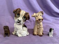 Four dog figurines displayed on purple fabric background: large ceramic dog with big green eyes, medium beige ceramic dog, small carved wooden dog, and tiny black and white ceramic dog.