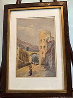 Framed watercolor painting showing the entire artwork including mat and frame, depicting a street with tall buildings, arch, and people walking.