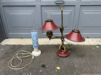 Photo showing both lamps outdoors on pavement in front of a garage door, one red brass-metal dual-shade lamp and one blue ceramic lamp with marble base.