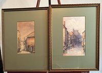 Framed pair of French watercolour paintings on easel showing two village scenes.