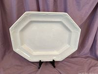Octagonal white ironstone porcelain platter with raised edge details displayed on a black stand against a purple draped background. Shows the front glossy glazed surface with minor glazing imperfections.