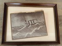 Framed black and white antique photo showing HMS Berwick in stormy ocean waves, wood frame and beige mat visible
