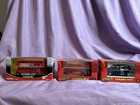 Three British model vehicles: two London buses and one London taxi, each in original packaging, placed in a row with a purple fabric background
