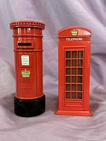 Two British-themed piggy banks: red post box and red phone booth, side by side on draped fabric background.