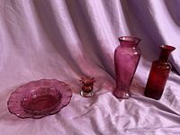 Four cranberry glass pieces shown grouped together on a lavender fabric backdrop: a round bowl, a small flower-shaped vase, and two tall vases of different shapes and sizes.