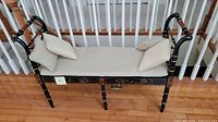 Front and side angle view of the Asian-inspired black wooden bench with cream cushion and two pillows on hardwood floor near staircase.