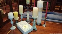 Photo showing various candleholders of different materials with pillar and taper candles arranged on wood table alongside a boxed set of Jerome Threewick candleholders.