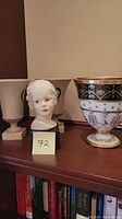 Porcelain bust of a child on black pedestal along with a decorative large vase on a wooden shelf.