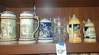 Six German beer steins and one clear glass beer mug placed on a wooden shelf. The steins have pewter lids (some), colorful relief and painted scenes.