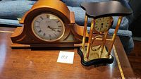 Two clocks placed side by side. The Seth Thomas wooden mantle clock measures approximately 17 inches in width and has a curved top. The Bey Berk desk clock has four gold-tone pillars and black top and base with a white clock face.