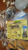 Assorted silverware including spoons, forks, knives, and small serving utensils arranged on a table with metal trays and some silverware in a yellow organizer tray.