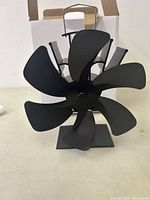 Photo showing a black metal stove fan with six blades sitting on its base, in front of its white box.