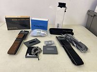 Photo showing all items in the lot arranged on a flat surface, includes beanie hat box, reading glasses, belts, chain bracelet, and metal item box