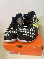 Front view of Nike Metcon 7 shoes, black and white mesh pattern with yellow swoosh, placed on orange Nike box showing minimal wear.