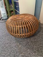 Brown wicker footstool on a carpeted floor, showing its circular shape and woven pattern.