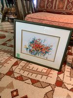 Framed floral painting resting on a carpet with geometric pattern, showing vibrant orange and blue flowers with green and yellow details. The frame is green with white mat and a thin gold line.
