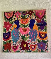 Frontal photo showing entire framed woven textile with birds and floral design against textured white wall.