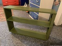 Photo showing the front and side angle of the green wood bookshelf on a carpeted floor near blue and red furniture items.