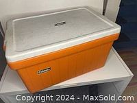 Orange plastic Coleman cooler with white lid on a white shelf, showing front and top sides.