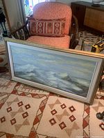 Framed signed mountain top landscape painting leaning against an upholstered chair, showing overall size and frame.