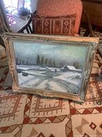 The full framed winter landscape painting resting on a chair on a patterned rug, showing snow-covered rural scene with trees and cottages.