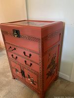 Front and side view showing the reddish-orange chest with black hand-painted designs of traditional Chinese figures and floral patterns, two drawers with metal handles, and bottom cabinet doors with a lock.