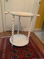Front angle view of the small white stand showing twisted spindle legs and circular shelves on a patterned rug over hardwood floor.