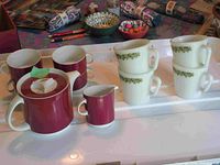 Wide view of two tea sets including Pyrex and Thomas with cups and milk jug in green and white and red and white colors.