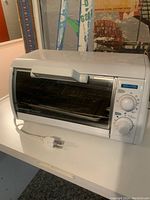 White plug-in toaster oven with pull-down front glass door, visible control dials, and power cord on a white surface.