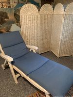 White plastic lounge chair with blue cushions shown fully extended indoors on carpeted floor with wicker room divider in background.