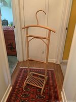 Front view of the wooden valet stand showing the hanger top, horizontal bar, slatted bottom shelf, and overall structure on a patterned rug.