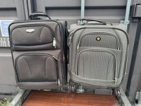 Two carry on suitcases, one dark gray and one light gray, placed side by side on a wooden surface in front of a metal wall.