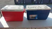 Two coolers on a white table outdoors. Left cooler is red with white lid; right cooler is blue with white lid and spray painted.
