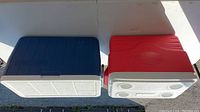 Top view of two rectangular Coleman coolers, one blue and one red, resting on a flat surface showing their lids and general shape.