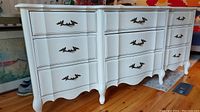 Front view of white solid wood dresser showing 9 drawers and ornate drawer handles.