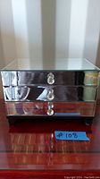 Front view of mirrored jewelry box showing three drawers with crystal knobs and black base on wood surface.