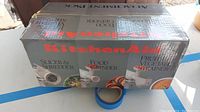 Box exterior showing KitchenAid branding and listing of included attachments: slicer & shredder, food grinder, fruit & vegetable strainer.