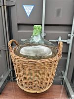 Large glass carboy inside woven wicker basket with two handles, standing upright on wooden surface with container door backdrop