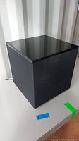 Black cube-shaped subwoofer with fabric sides and glossy top surface, shown from a front corner angle on a white surface.